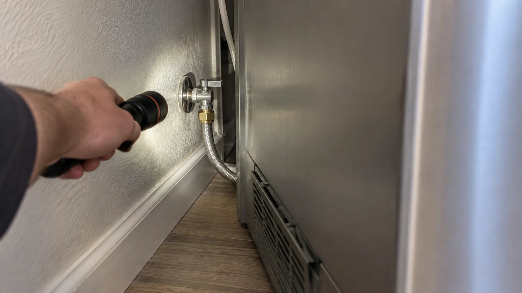 Close on a flashlight lighting the supply line behind a new refrigerator.