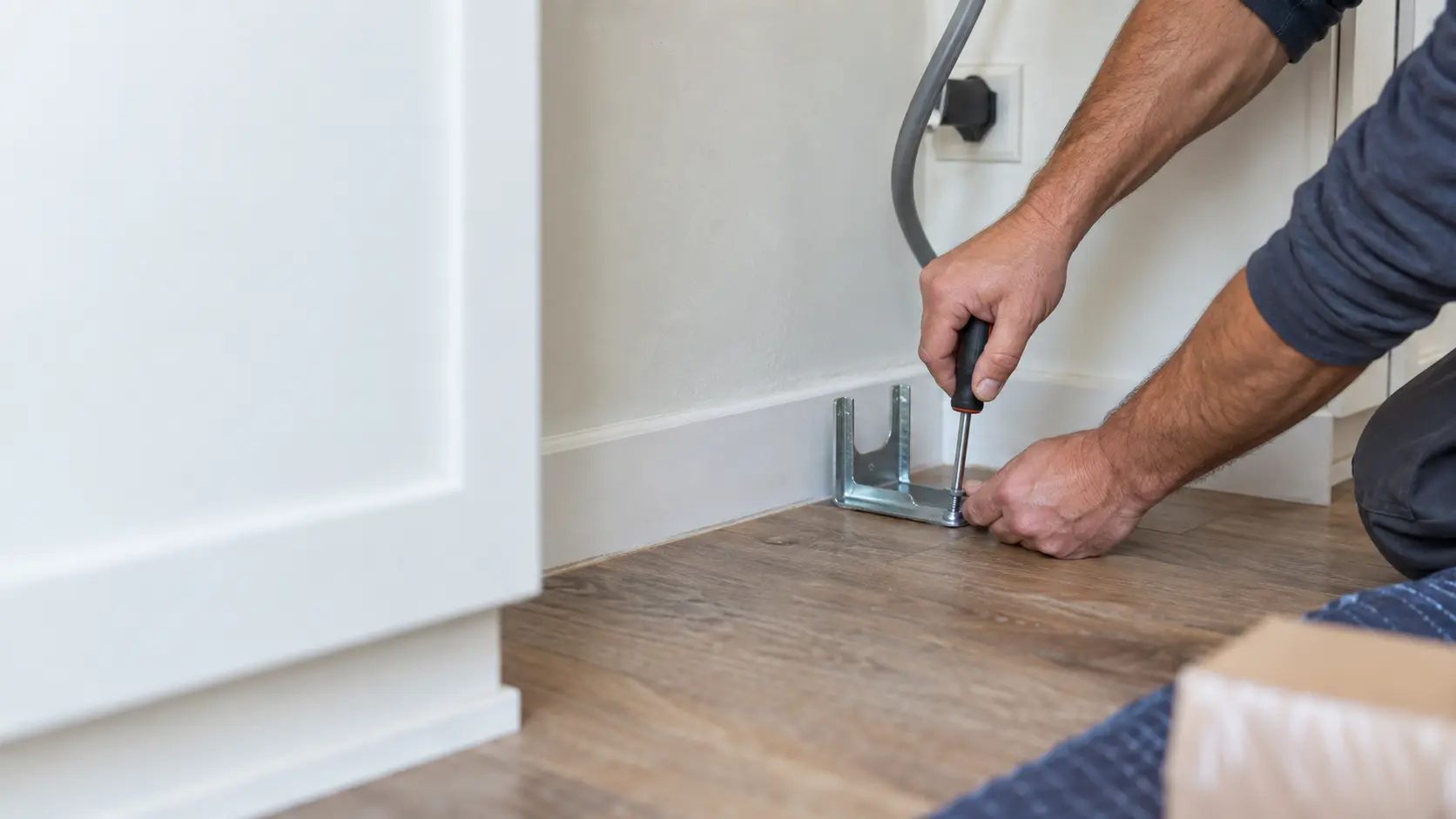 A technician's hands screwing an anti-tip bracket to the floor behind a range space.